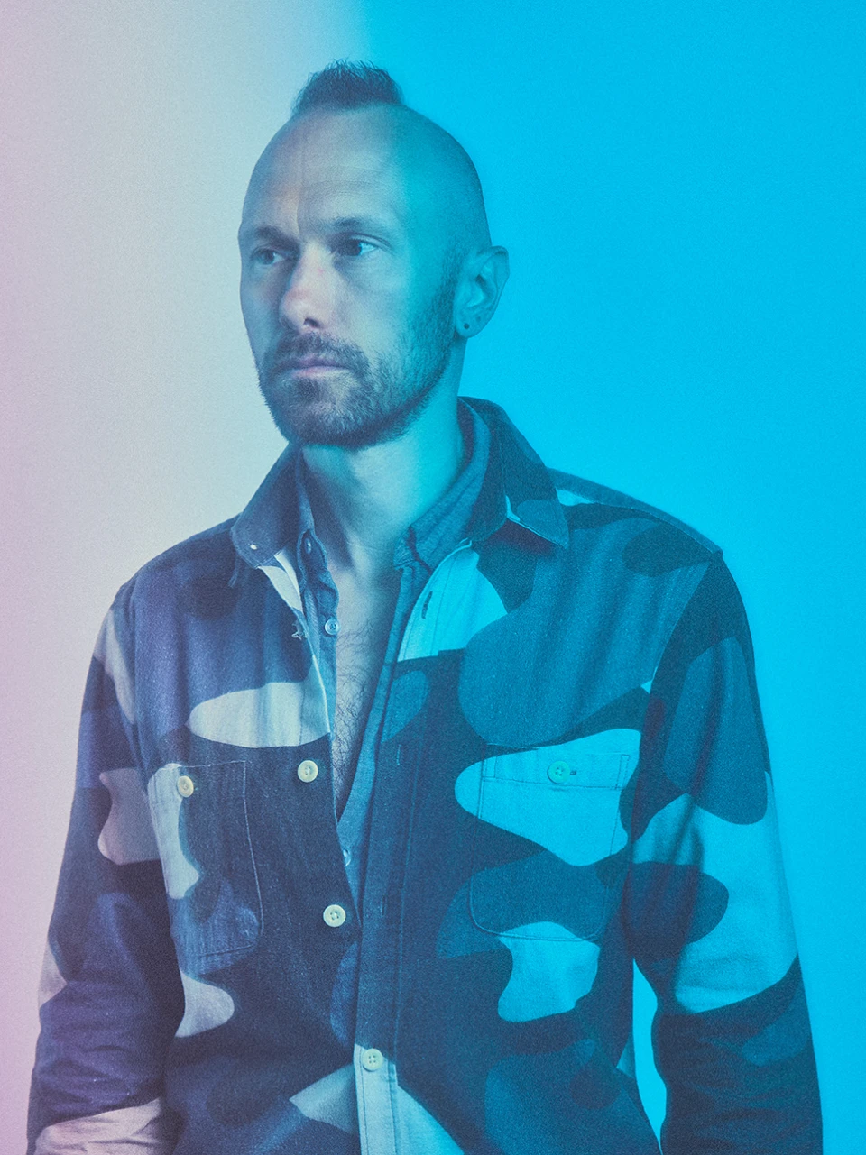 East Forest, musician and creator of immersive soundscapes, wearing a patterned shirt, against a gradient blue background, reflecting themes of spirituality and introspection.